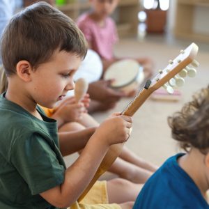 Nursery school children sitting on floor indoors in classroom, playing musical instruments.